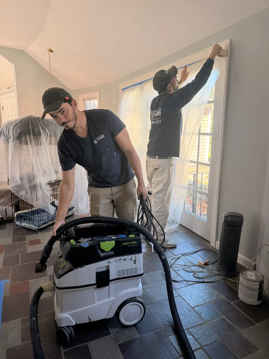 Two painters masking a room with plastic while a dust extractor vacuum is connected to the sander
